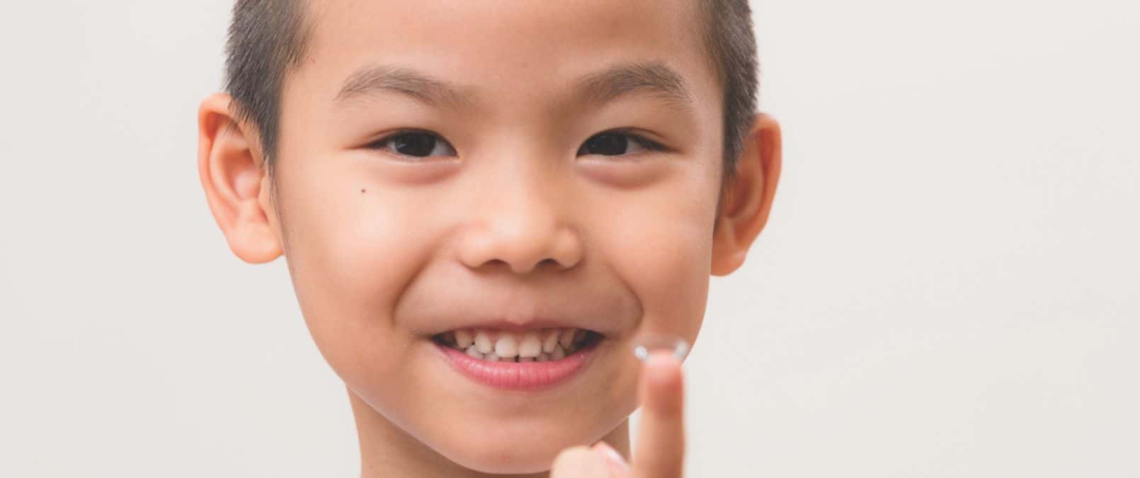 Close-up photograph of a smiling child holding a contact lens on the tip of their finger. Childhood myopia remedies include specialty contact lenses.